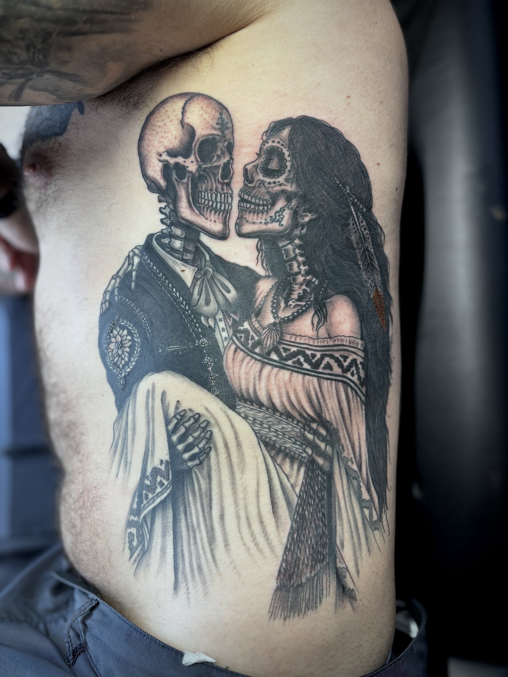 Black and grey tattoo of a skeleton couple embracing in traditional dress on the ribs